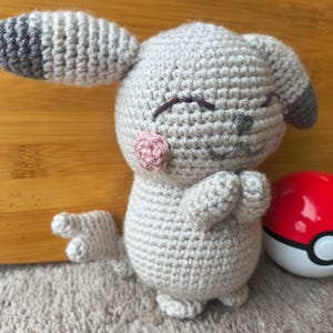 May include: A crocheted plush toy in the shape of a cartoon character, primarily light gray with darker gray and pink accents. The toy has closed eyes, pink cheeks, and a small tail. A red and white ball is next to the toy.