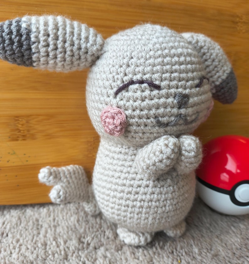 May include: A crocheted plush toy in the shape of a cartoon character, primarily light gray with darker gray and pink accents. The toy has closed eyes, pink cheeks, and a small tail. A red and white ball is next to the toy.