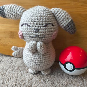 May include: A handmade crochet plush toy in the shape of a cartoon character, with gray yarn, pink cheeks, and closed eyes. A red and white Pok&eacute; Ball sits next to the plush toy. The toy has folded hands and gray and black ears.
