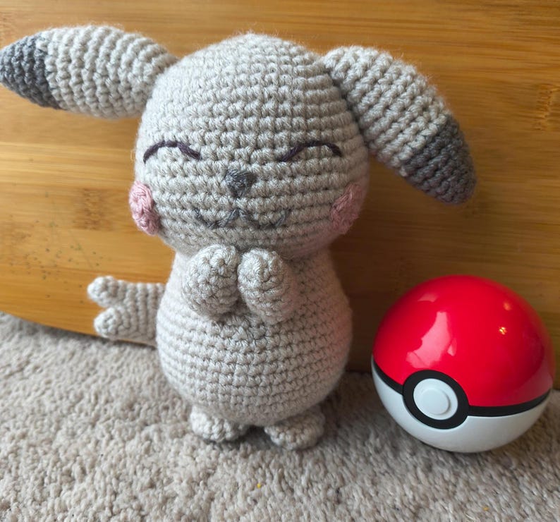 May include: A handmade crochet plush toy in the shape of a cartoon character, with gray yarn, pink cheeks, and closed eyes. A red and white Pok&eacute; Ball sits next to the plush toy. The toy has folded hands and gray and black ears.