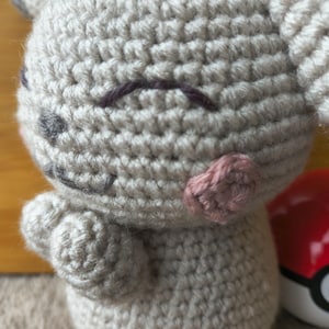 May include: A handmade crochet plush toy in light gray, featuring a smiling face with dark purple eyebrows, gray eyes, and pink cheeks. The toy has small arms and ears. A red and white ball is partially visible in the background.