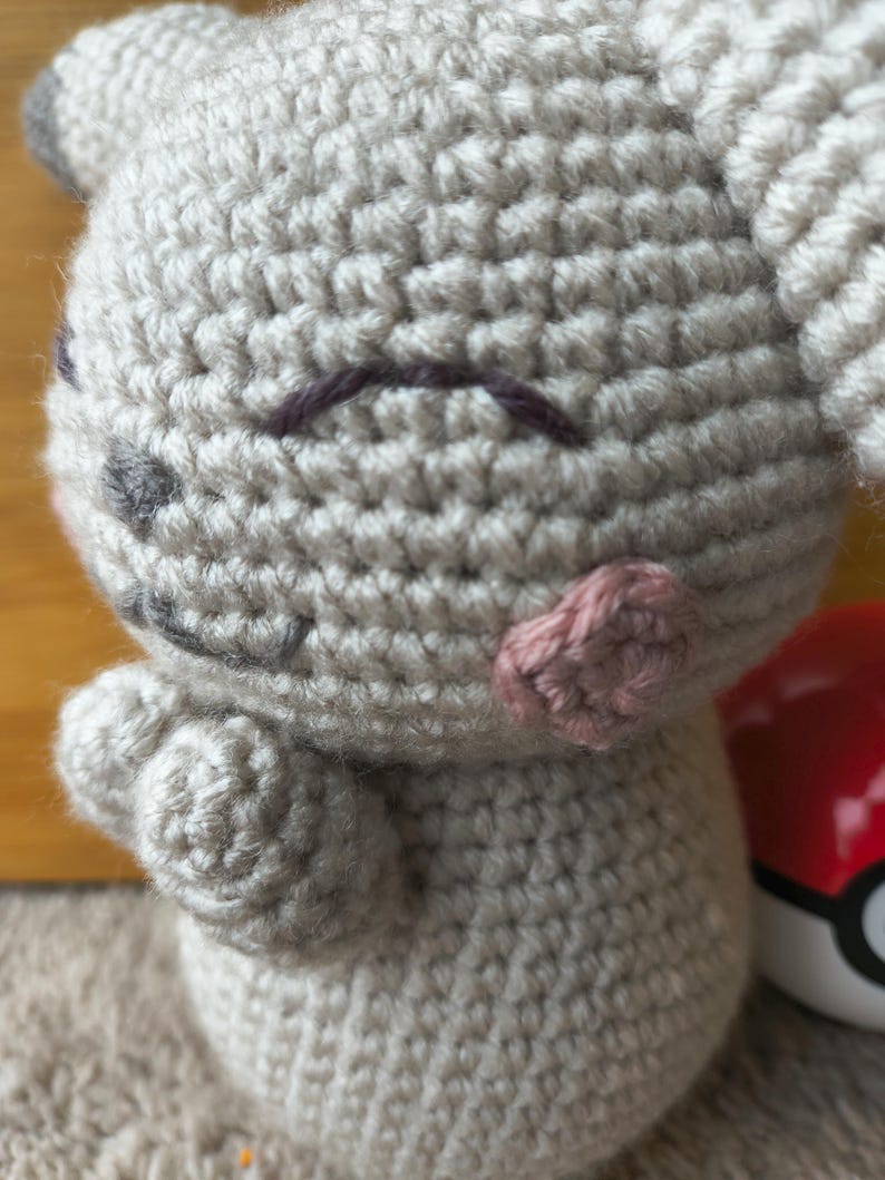 May include: A handmade crochet plush toy in light gray, featuring a smiling face with dark purple eyebrows, gray eyes, and pink cheeks. The toy has small arms and ears. A red and white ball is partially visible in the background.