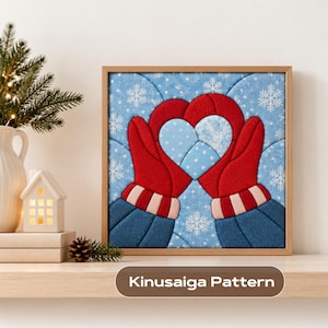 May include: A square framed Kinusaiga pattern art piece featuring two red mittens forming a heart shape against a light blue background with white snowflakes. The mittens have striped cuffs and are set in a light wood frame.