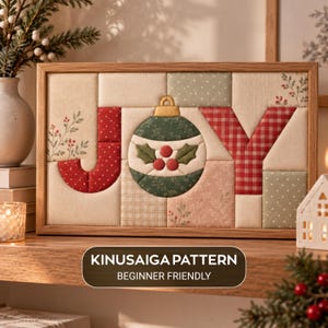 May include: A rectangular framed fabric art piece featuring the word JOY, where the letter O is replaced by a green Christmas ornament with holly berries. The design uses various patterned fabric patches in red, green, and cream. Text reads KINUSAIGA PATTERN BEGINNER FRIENDLY.