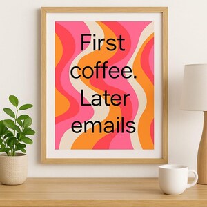 Op de afbeelding: Ingelijste print met een retro design in roze, oranje en crème. Het kunstwerk bevat de tekst "First coffee. Later emails" in zwart. De print wordt weergegeven op een houten tafel.