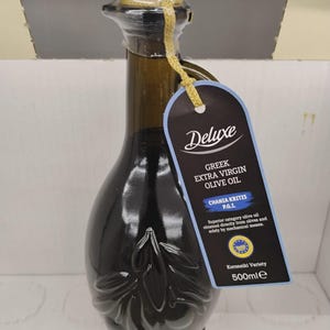 Puede incluir: Una botella de vidrio verde oscuro de aceite de oliva virgen extra griego con tapón de corcho y etiqueta decorativa. La botella tiene un diseño floral y una cuerda dorada. La etiqueta dice "Deluxe Greek Extra Virgin Olive Oil" y contiene 500 ml.