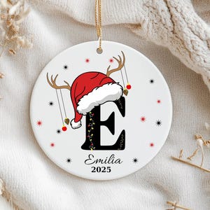 May include: A round white ceramic ornament featuring a large black letter E topped with a red Santa hat and reindeer antlers. The letter is decorated with colourful string lights. Below, the name Emilia and the year 2025 are printed in black.