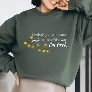 May include: Dark green sweatshirt featuring the text "Probably just gonna jingle some of the way, I'm tired." A string of colorful Christmas lights is featured below the text. The sweatshirt is a casual, comfortable style.