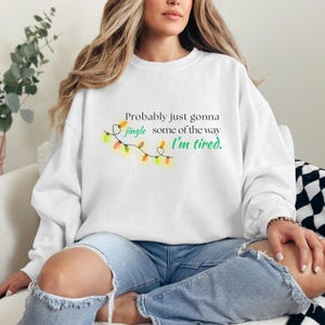 May include: White crew neck sweatshirt featuring the text "Probably just gonna jingle some of the way, I'm tired" and a string of Christmas lights. The design is in green, orange, and yellow.