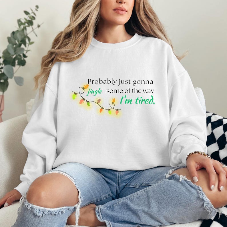 May include: White crew neck sweatshirt featuring the text "Probably just gonna jingle some of the way, I'm tired" and a string of Christmas lights. The design is in green, orange, and yellow.