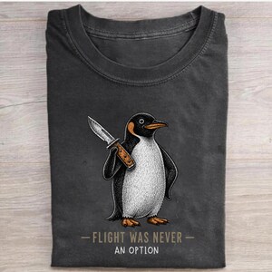 May include: A charcoal grey crew neck t-shirt featuring a graphic illustration of a penguin holding a knife. Below the penguin, the text reads FLIGHT WAS NEVER AN OPTION in a simple, light-coloured font. The shirt has a classic short-sleeve design.