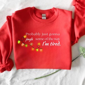 May include: A red crew neck sweatshirt with the text "Probably just gonna jingle some of the way I'm tired." A string of colourful Christmas lights is featured on the left side of the text. The sweatshirt is made of a soft material.