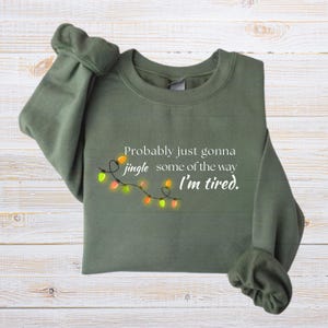 May include: An olive green sweatshirt featuring the text "Probably just gonna jingle some of the way I'm tired." A string of colourful Christmas lights is below the text. The sweatshirt is folded on a light wood surface.