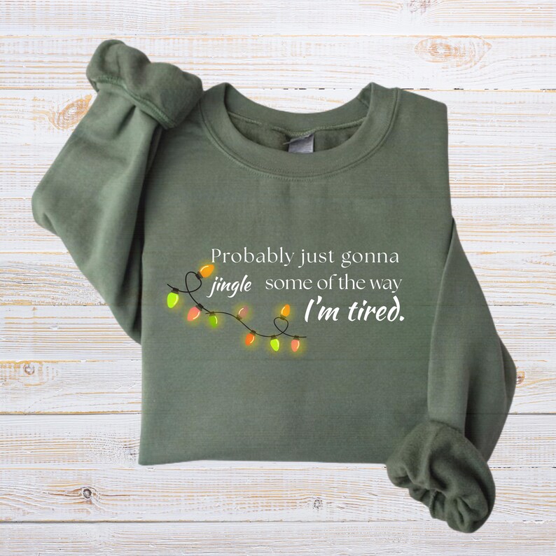 May include: An olive green sweatshirt featuring the text "Probably just gonna jingle some of the way I'm tired." A string of colourful Christmas lights is below the text. The sweatshirt is folded on a light wood surface.