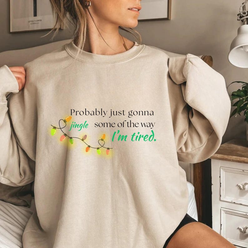 May include: Beige sweatshirt featuring the text "Probably just gonna jingle some of the way I'm tired." The text is in various colours, with a string of Christmas lights. The sweatshirt is a neutral colour.