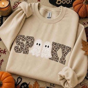 May include: A beige crewneck sweatshirt featuring the word SPOOKY in block letters with a brown and black leopard print pattern. Two white lace ghost figures with black eyes are positioned in the center, replacing the letter O.