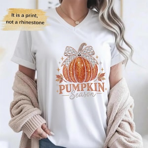 May include: A white V-neck t-shirt featuring a graphic of an orange pumpkin with a plaid bow and the text "PUMPKIN Season". The design is a print, not a rhinestone. The shirt has short sleeves and is worn with a beige cardigan.