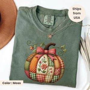 May include: A moss green t-shirt featuring a graphic of a patchwork pumpkin with various patterns including polka dots, gingham, and plaid. The pumpkin is decorated with a large red bow and floral accents. A silver necklace rests on the neckline.