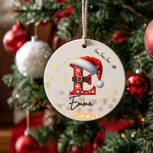 May include: A round Christmas ornament with a festive design. The ornament features a red letter "E" adorned with snowflakes and a Santa hat. The name "Emma" is written below the letter in cursive. A string is attached for hanging.