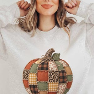 May include: A light gray crewneck sweatshirt featuring a large, central graphic of a pumpkin constructed from a patchwork of various fabric patterns, including leopard print, black and brown buffalo plaid, yellow gingham, and floral prints.