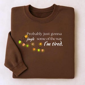 May include: A brown sweatshirt featuring the phrase "Probably just gonna jingle some of the way I'm tired." The design includes a string of festive lights. The garment is neatly folded.