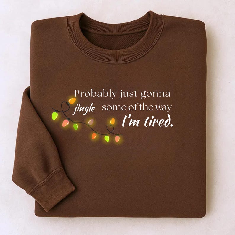May include: A brown sweatshirt featuring the phrase "Probably just gonna jingle some of the way I'm tired." The design includes a string of festive lights. The garment is neatly folded.