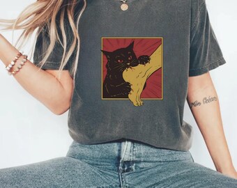 May include: Dark grey t-shirt featuring a graphic of a black cat biting a leg. The cat has red eyes and is set against a red and gold background. The t-shirt is a casual style.