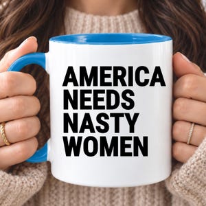 May include: A white ceramic mug with a bright blue handle and interior rim. The front of the mug features bold, black, sans-serif text that reads: AMERICA NEEDS NASTY WOMEN, arranged in four stacked lines of capitalized lettering.