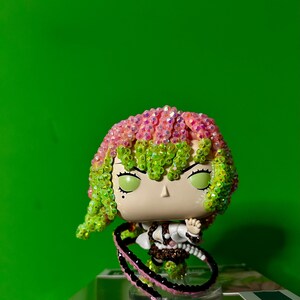 May include: A Funko Pop figure of a character with pink and green hair, green eyes, and a determined expression. The figure is wearing a white and brown outfit and has a black and pink whip. The background is a solid green.