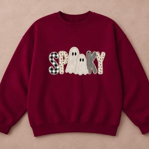 Puede incluir: Una sudadera de cuello redondo color granate con la palabra SPOOKY impresa en el pecho. Las letras están rellenas con varios patrones, incluyendo cuadros negros y blancos, rayas verticales negras y blancas y un estampado floral, con dos fantasmas.