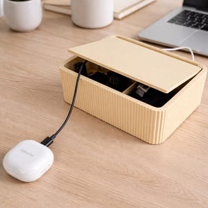 Puede incluir: Un organizador de cables rectangular beige con exterior acanalado y tapa con bisagras. Un dispositivo de carga Samsung blanco está conectado al organizador con un cable negro. El organizador está sobre una superficie de madera clara.