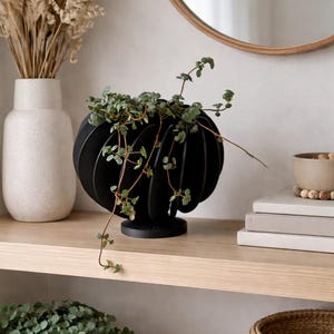 Op de afbeelding: Een zwarte, bolvormige plantenbak met een uniek, gelaagd ontwerp, met een hangende groene plant. De plantenbak staat op een houten plank, samen met een witte vaas met gedroogde grassen, een spiegel en andere decoratieve items. Woondecoratie.