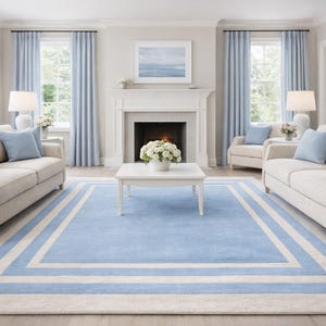 Könnte beinhalten: Ein lichtdurchflutetes Wohnzimmer mit neutralfarbenen Sofas, blauen Zierkissen und passenden Vorhängen. Ein blau-weiß gemusterter Teppich bildet den Mittelpunkt des Raumes, ergänzt durch einen weißen Couchtisch und einen Kamin. Der Raum ist klassisch eingerichtet.