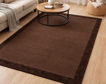 May include: A rectangular area rug featuring a solid dark brown centre with a textured, grid-like pattern and a smooth, wide border in a matching dark brown tone. The rug is positioned flat on a light-coloured wood floor, providing a neutral foundation.