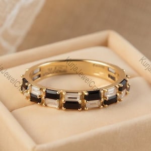 Puede incluir: Un anillo de banda dorado con piedras rectangulares negras y blancas alternas. El anillo está colocado en una caja de terciopelo beige. El nombre de la marca "KurjaJewelCraft" es visible en el fondo.