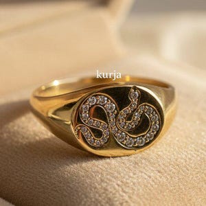 Könnte beinhalten: Vergoldeter Siegelring mit Schlangenmuster, besetzt mit kleinen, funkelnden Steinen. Der Ring wird in einer beigen Schmuckschatulle präsentiert. Das Wort "kurja" ist auf dem Ring sichtbar.