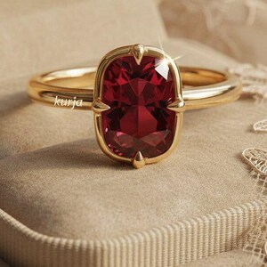 May include: A gold ring featuring a large, cushion-cut deep red gemstone held by four prongs. The band is smooth and polished gold. The word kurja is printed in small white letters on the side of the ring band near the central gemstone setting.