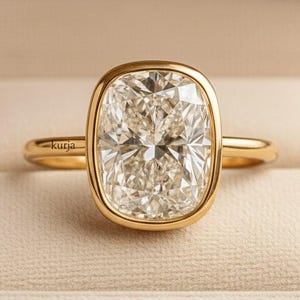 Puede incluir: Un anillo de oro con una gema grande, rectangular y transparente. La gema está engastada en un bisel. El anillo está en un joyero beige. La palabra "kurja" está grabada en el anillo.