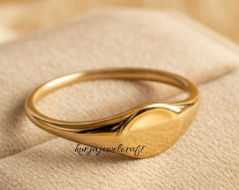 May include: A gold signet ring with a smooth, oval face. The ring is displayed on a beige velvet surface inside a jewelry box. The ring has the text "kurjajewelcraft" engraved on the side.