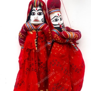 May include: Two traditional Indian puppets. The puppets are dressed in red fabric with gold accents. They have white painted faces with detailed features and are suspended by red strings.