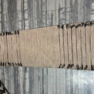 May include: A rectangular woven table runner in a natural beige tone featuring a textured fabric construction. The runner is accented with vertical black thread stripes and decorative black fringe tassels along both of the shorter ends.