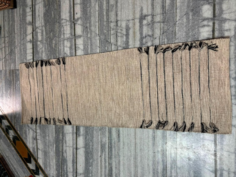 May include: A rectangular woven table runner in a natural beige tone featuring a textured fabric construction. The runner is accented with vertical black thread stripes and decorative black fringe tassels along both of the shorter ends.