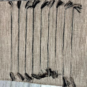 May include: A rectangular woven table runner in a neutral beige tone featuring vertical black stripes. The runner is accented with decorative black and beige tassels attached at both the top and bottom edges, creating a textured appearance.