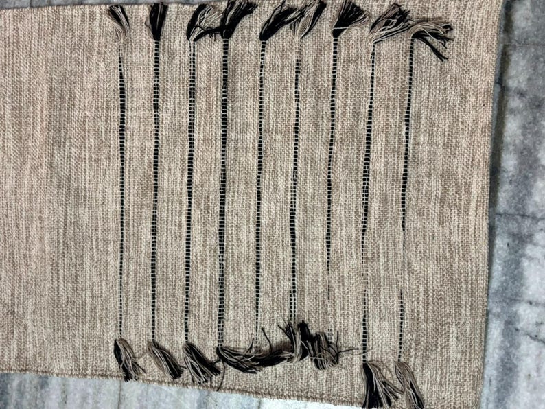 May include: A rectangular woven table runner in a neutral beige tone featuring vertical black stripes. The runner is accented with decorative black and beige tassels attached at both the top and bottom edges, creating a textured appearance.