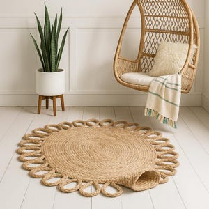 Puede incluir: Una alfombra redonda de yute natural con borde festoneado. La alfombra está hecha de fibras tejidas y es de color marrón claro. Está colocada sobre un suelo de madera blanco, con una silla colgante de mimbre y una planta en maceta en el fondo.