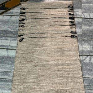 May include: A rectangular woven runner rug featuring a textured beige and tan surface with horizontal black stripes. The rug is finished with small, decorative black and tan tassels attached along the two long sides and at the corners.