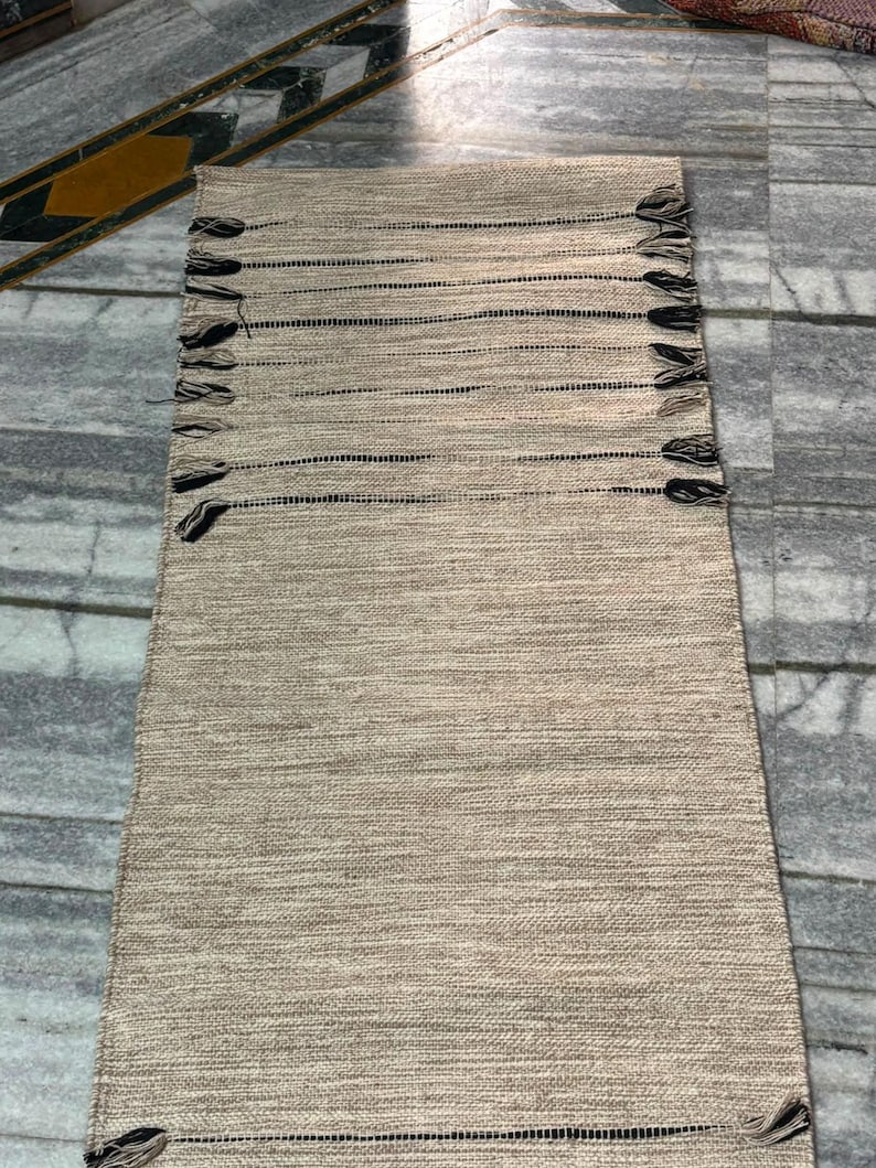 May include: A rectangular woven runner rug featuring a textured beige and tan surface with horizontal black stripes. The rug is finished with small, decorative black and tan tassels attached along the two long sides and at the corners.
