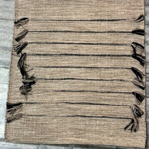 May include: A rectangular table runner featuring a woven tan fabric with a series of horizontal black stripes. The runner is accented with decorative tassels in alternating black and tan colors attached along the side edges of the fabric.