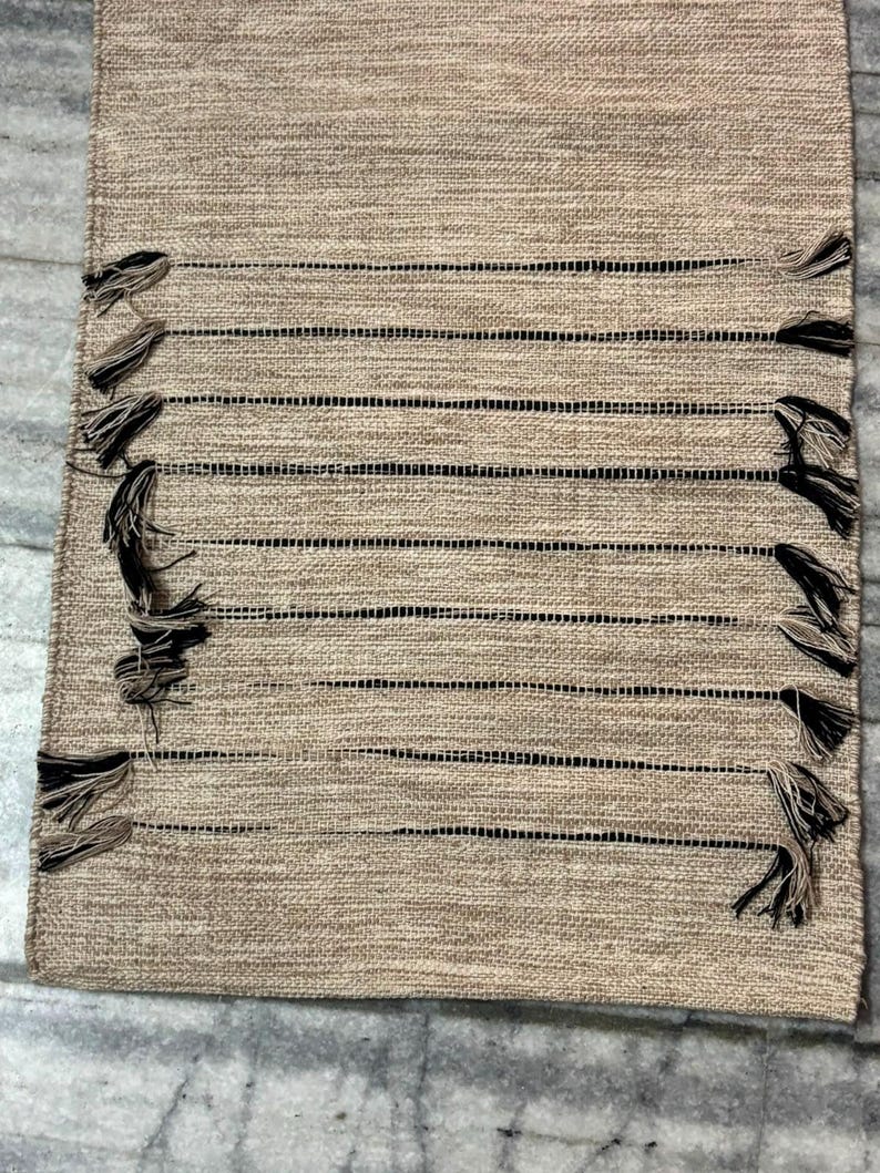 May include: A rectangular table runner featuring a woven tan fabric with a series of horizontal black stripes. The runner is accented with decorative tassels in alternating black and tan colors attached along the side edges of the fabric.