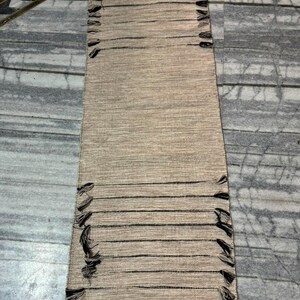 May include: A rectangular woven table runner in a natural beige hue, featuring horizontal dark brown thread accents and decorative fringe detailing along the side edges. The runner has a textured, fibrous appearance with a consistent weave pattern.